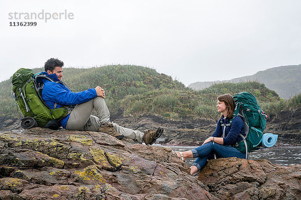 Junges Paar mit Rucksack  auf Felsen sitzend  Constant Bay  Charleston  Südinsel  Neuseeland
