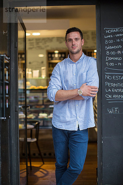 Porträt eines mittelgroßen  männlichen Cafébesitzers  der sich an die Café-Tür lehnt