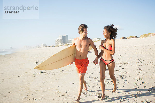 Junges Surferpaar läuft Hand in Hand am Strand  Kapstadt  Western Cape  Südafrika