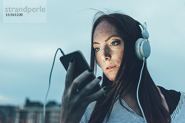 Junge sommersprossige Frau hört Kopfhörer und schaut in der Abenddämmerung auf das Smartphone.