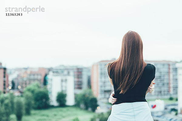 Rückansicht einer jungen Frau mit langen roten Haaren  die über das Stadtbild blickt.