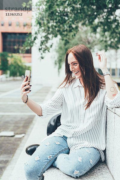 Junge Frau sitzt an der Wand und nimmt Smartphone Selfie