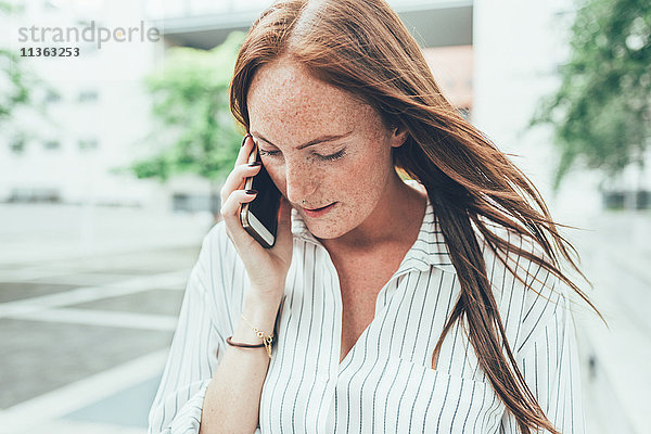 Junge Frau mit Sommersprossen und langen roten Haaren macht Smartphone-Anruf vor dem Bürogebäude