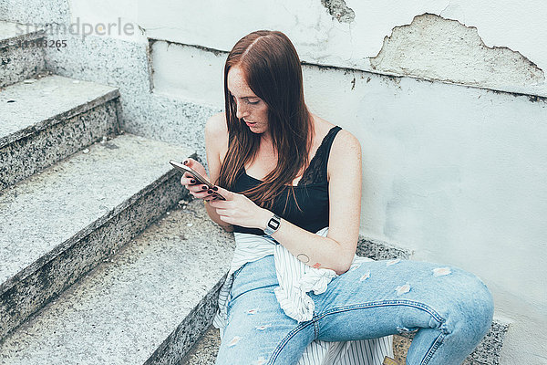 Junge Frau sitzt auf der Treppe und liest Smartphone-Texte.