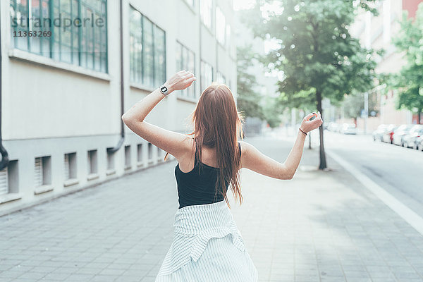 Rückansicht einer jungen Frau mit langen roten Haaren  die auf der Straße tanzt.
