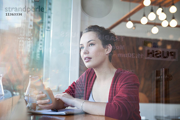 Junge Frau sitzt im Café  hält ein Smartphone in der Hand und schaut aus dem Fenster