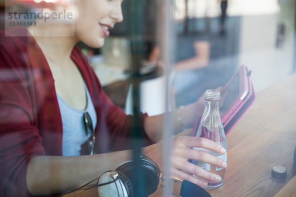Junge Frau sitzt im Café  hält eine Flasche Wasser in der Hand und benutzt ein digitales Tablett