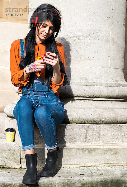 Junge Frau sitzt neben einer Säule  trägt Kopfhörer und hält ein Smartphone