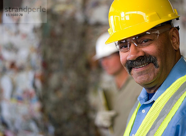 Porträt eines reifen männlichen Arbeiters mit Schutzhelm  vor gestapeltem Müll in der Recycling-Anlage