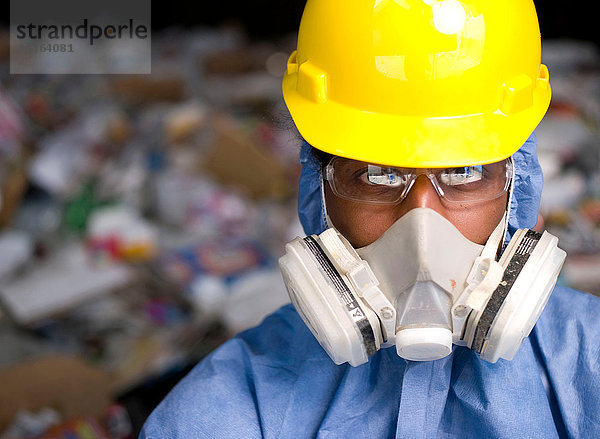 Porträt eines männlichen Arbeiters mit Schutzhelm und Gasmaske  vor dem Müll der Recycling-Anlage
