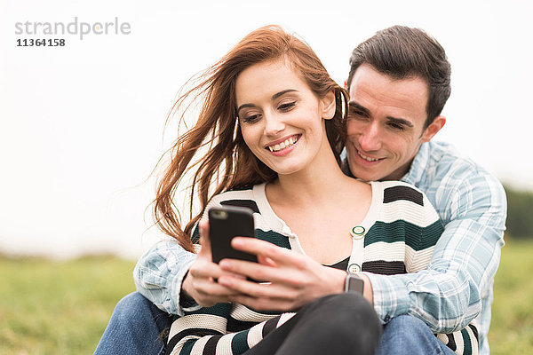 Ein Paar sitzt im Feld und schaut auf das Smartphone.