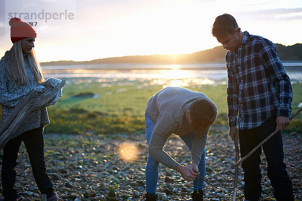 Drei erwachsene Freunde sammeln bei Sonnenuntergang Treibholz im Meer