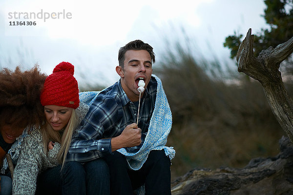 Junger Mann und Freundin essen getoastete Marshmallows am Strand in der Abenddämmerung