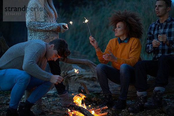 Vier junge erwachsene Freunde spielen mit Wunderkerzen am Lagerfeuer am Strand in der Abenddämmerung