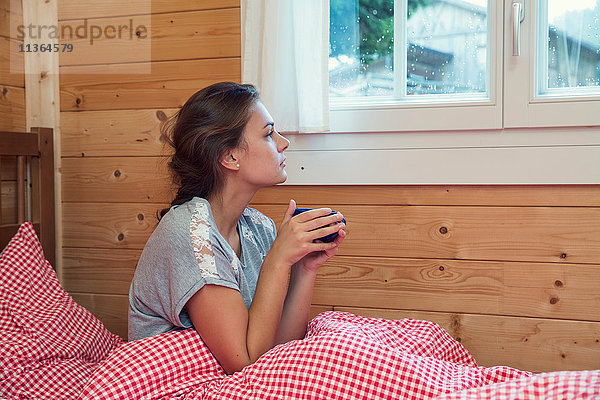 Junge Frau schaut durchs Fenster vom Blockhausbett  Tirol  Österreich