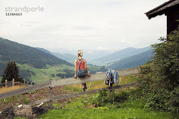 Rückansicht einer auf einem Zaun sitzenden Frau mit Blick auf die Berglandschaft  Sattelbergalm  Tirol  Österreich