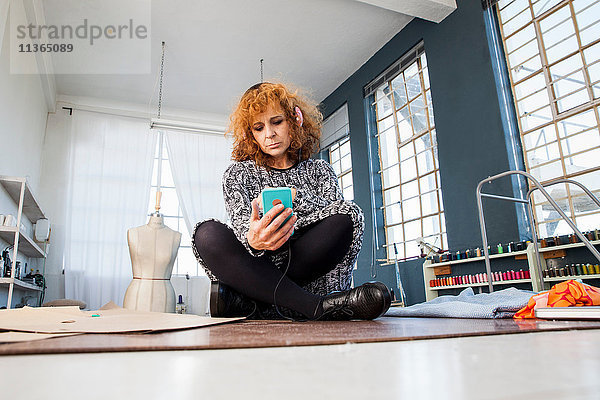 Reife Modedesignerin im Schneidersitz auf dem Werkstatttisch mit Blick auf das Smartphone