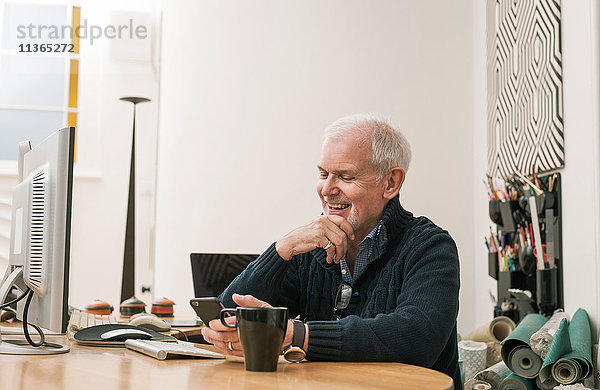 Älterer Mann lächelt  benutzt Mobiltelefon am Arbeitstisch