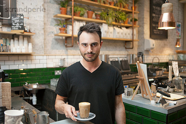 Porträt eines jungen männlichen Barista  der in einem Café ein Glas Kaffee serviert
