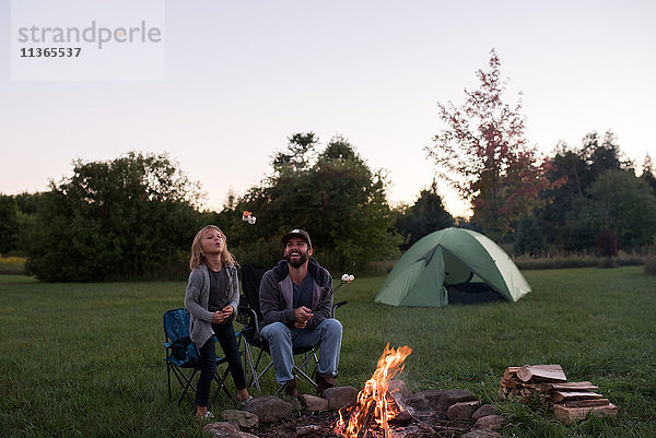 Vater und Tochter sitzen am Lagerfeuer und rösten Marshmallows über dem Feuer
