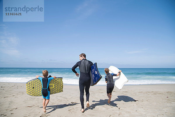 Rückansicht eines Mannes und zweier Söhne  die mit Bodyboards am Strand entlang rennen  Laguna Beach  Kalifornien  USA