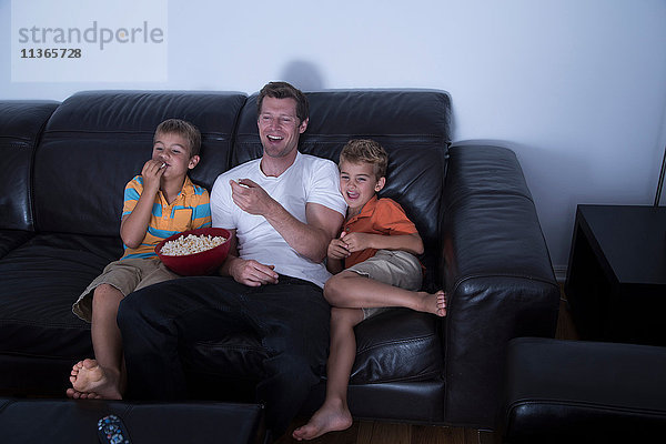 Mann und zwei Söhne lachen vor dem Fernseher  während sie auf dem Sofa Popcorn essen