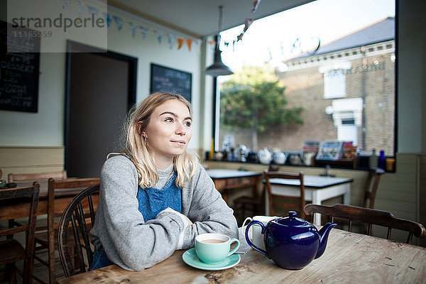 Junge Frau im Café sitzend  Tasse Tee und Teekanne auf dem Tisch  nachdenklicher Ausdruck