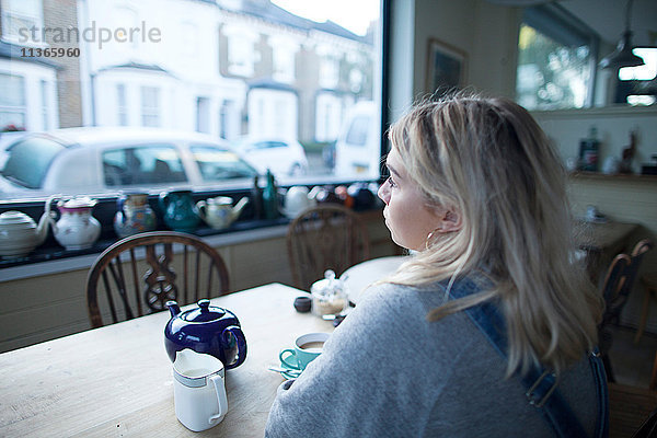 Junge Frau im Café sitzend  Tasse Tee und Teekanne auf dem Tisch  Blick aus dem Fenster