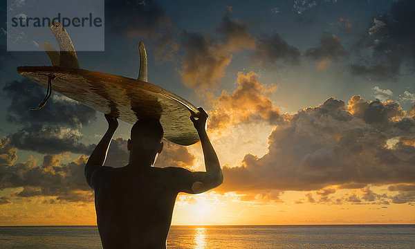 Silhouettierte Rückansicht eines männlichen Surfer  der ein Surfbrett auf dem Kopf trägt und den Sonnenuntergang über dem Meer beobachtet  Rio De Janeiro  Brasilien