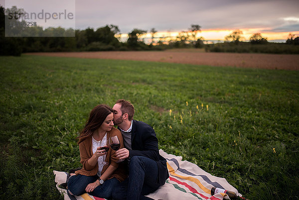 Romantisches Paar mit Rotwein  das sich bei Sonnenuntergang auf einer Picknickdecke im Feld entspannt