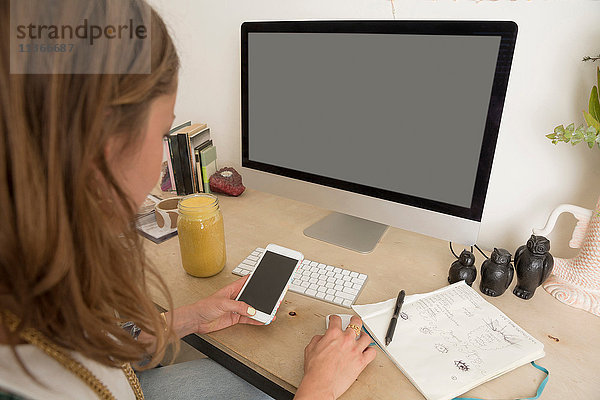 Frau sitzt am Schreibtisch und benutzt Computer und Smartphone