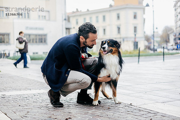 Mittelgroßer erwachsener Mann  der auf einem gepflasterten Stadtplatz kauert  um seinen Hund zu streicheln