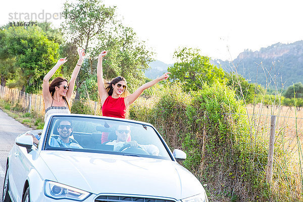 Zwei junge Freundinnen stehen und winken  während sie mit einem Cabrio auf einer Landstraße fahren  Mallorca  Spanien