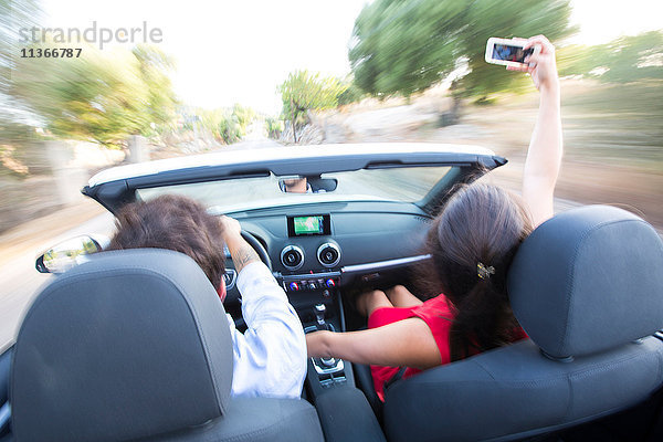 Rückansicht eines jungen Paares  das sich beim Fahren auf einer Landstraße im Cabriolet selbstständig macht  Mallorca  Spanien