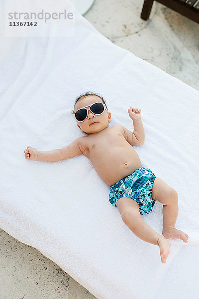 Baby mit Sonnenbrille auf Matratze liegend