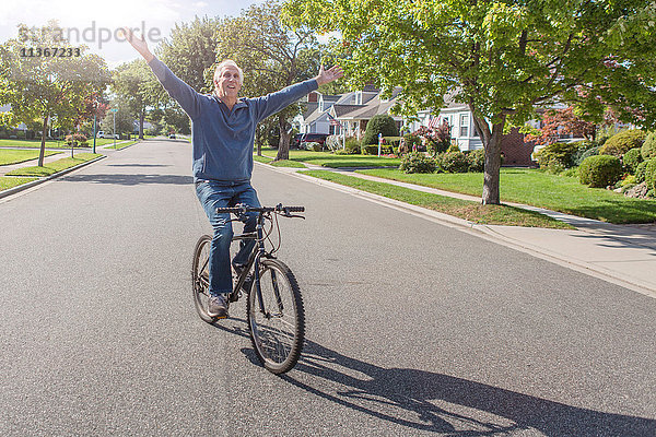 Älterer Mann mit erhobenen Händen beim Radfahren entlang einer Vorstadtstraße