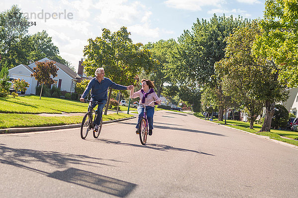 Romantisches älteres Ehepaar hält sich an der Hand und fährt mit dem Fahrrad die Vorstadtstraße entlang