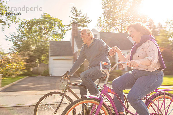 Romantisches älteres Ehepaar hält sich an der Hand und fährt mit dem Fahrrad die Vorstadtstraße entlang