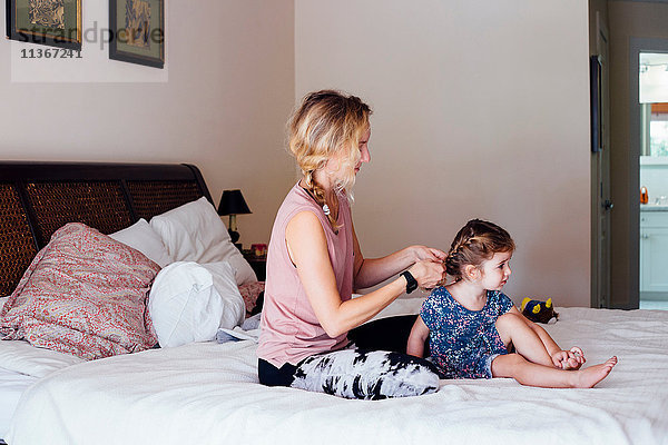 Frau sitzt auf dem Bett und flechtet die Haare der Kleinkind-Tochter
