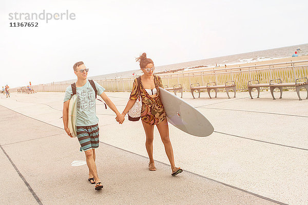 Junges Paar hält Hände und trägt Surfbretter am Rockaway Beach  New York State  USA