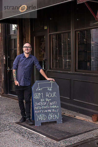 Porträt eines Barmanns  der sich vor dem Fenster einer Kneipe an ein Happy-Hour-Brett lehnt  Brooklyn  New York  USA