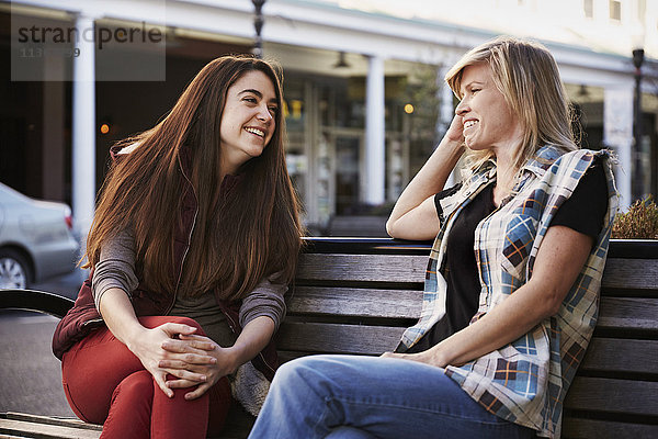 Zwei Frauen sitzen auf einer Bank in einer städtischen Umgebung und unterhalten sich.