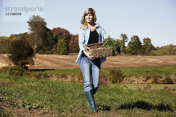Eine junge Frau in Arbeitskleidung geht über ein Feld und hält einen Korb mit Feldfrüchten in der Hand.