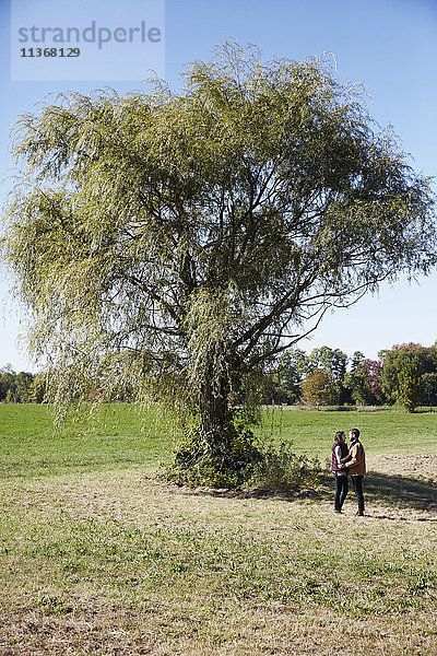 Eine junge Frau und ein junger Mann stehen neben einem Baum auf einem Feld und schauen sich gegenseitig an.