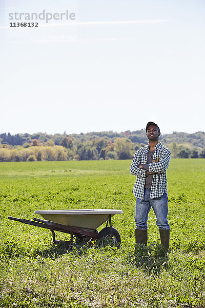 Ein junger Mann in Arbeitskleidung steht in einem Getreidefeld mit verschränkten Armen neben einer Schubkarre.