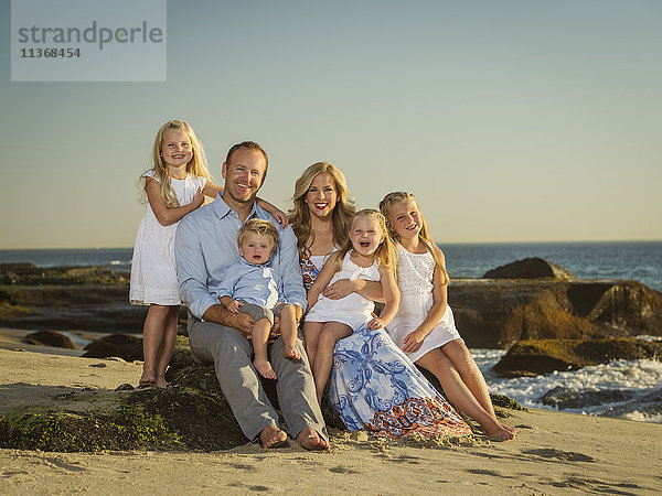Familie mit Kindern (12-17 Monate  4-5  6-7  8-9) sitzend am Strand mit Meer im Hintergrund