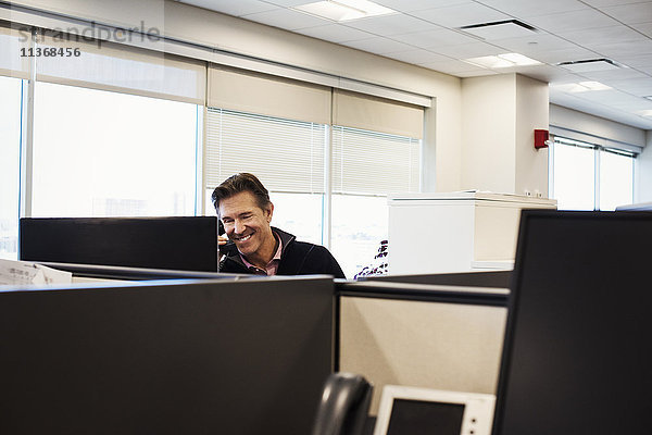Ein Mann sitzt in einer Bürokabine mit einem Telefon am Ohr und lächelt.