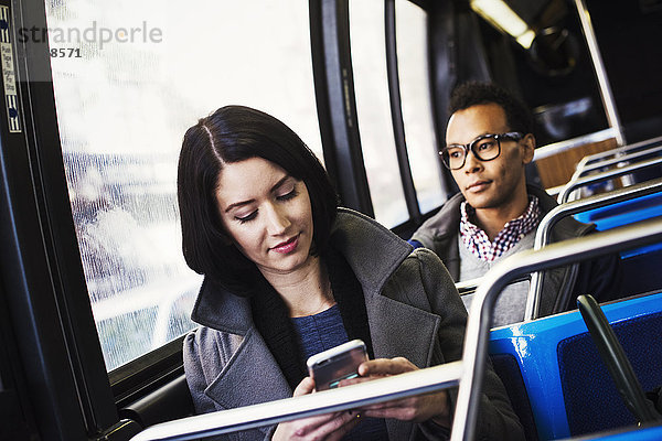 Eine junge Frau und ein junger Mann sitzen in öffentlichen Verkehrsmitteln  einer schaut auf ein Handy und einer schaut weg.