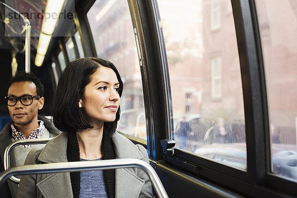 Eine junge Frau sitzt in einem Zug und blickt aus dem Fenster auf eine Stadtlandschaft  hinter ihr sitzt ein Mann und schaut weg.