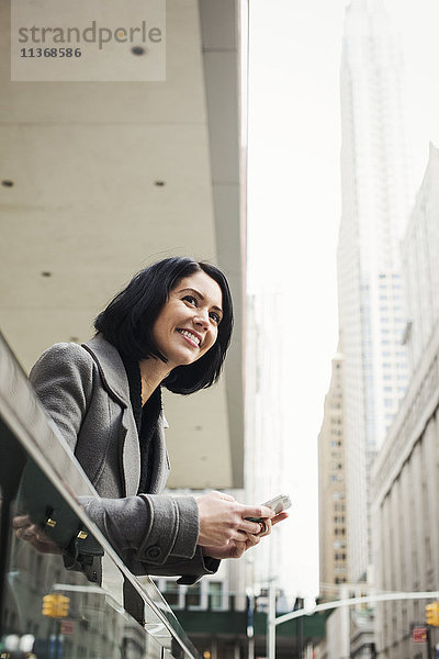 Eine junge Frau lehnt lächelnd über einen Balkon und hält ein Handy in der Hand.